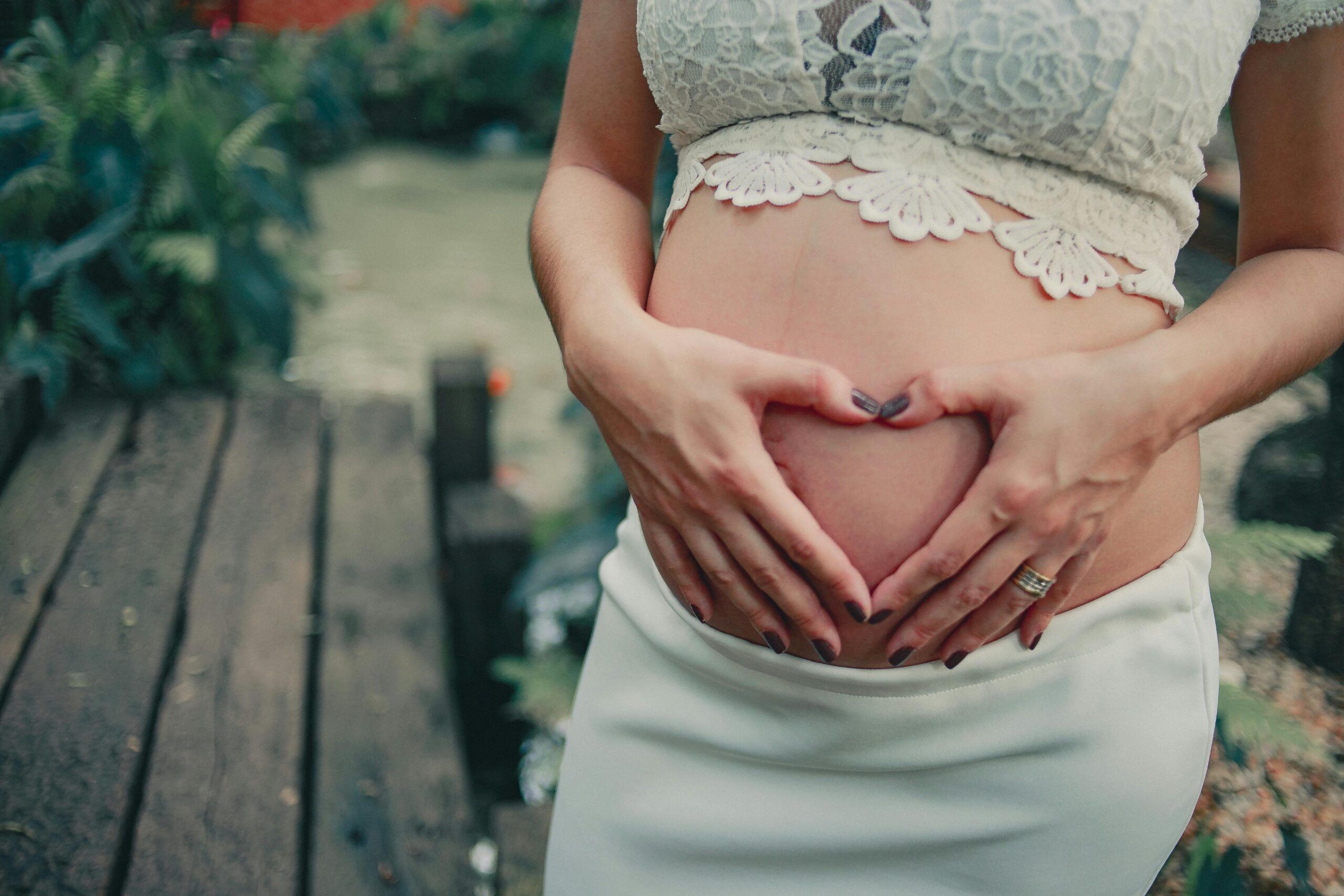 A pregnant woman making a heart shape on her baby bump in an outdoor setting.